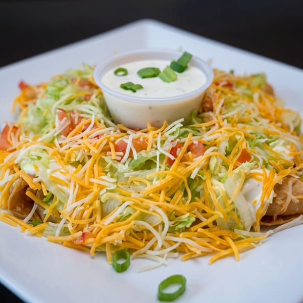 Crispy taquitos covered in shredded lettuce, cheese, diced tomatoes, and a side of ranch dressing.