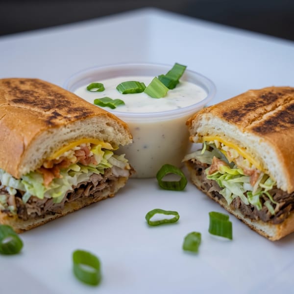 Two halves of a toasted sub sandwich with meat, cheese, and lettuce, next to a dipping sauce.