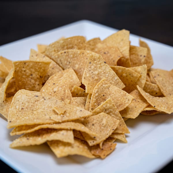 A white rectangular plate piled high with golden, triangular tortilla chips speckled with black pepper.